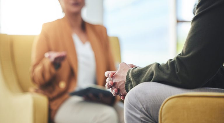A blurred, soft-focus view of a practitioner and a client seated in a modern, welcoming alcohol recovery centre, engaged in a supportive conversation.