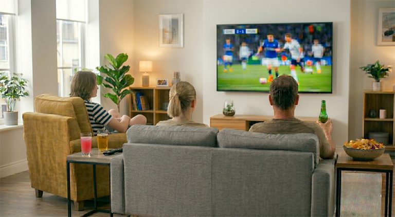 Two teenagers and their Dad sit watching football in a living room. There are drinks and snacks in the room.