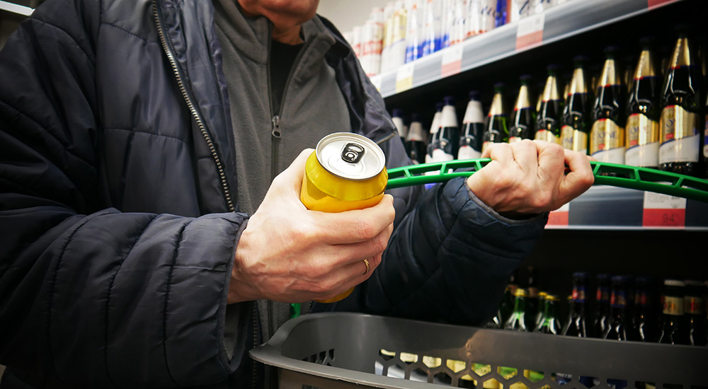 A man stands in a shop by a shelf full of bottles of alcohol. He holds a can of beer and a shopping basket in his hands.