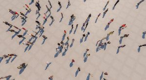An aerial, top-down photograph of numerous people walking across a light-coloured pavement, casting long distinct shadows. The image captures a diverse flow of pedestrians moving in various directions.
