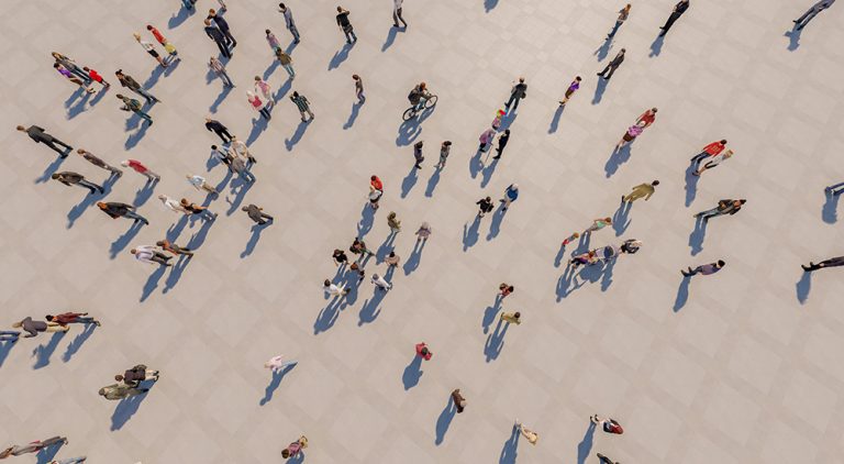 An aerial, top-down photograph of numerous people walking across a light-coloured pavement, casting long distinct shadows. The image captures a diverse flow of pedestrians moving in various directions.