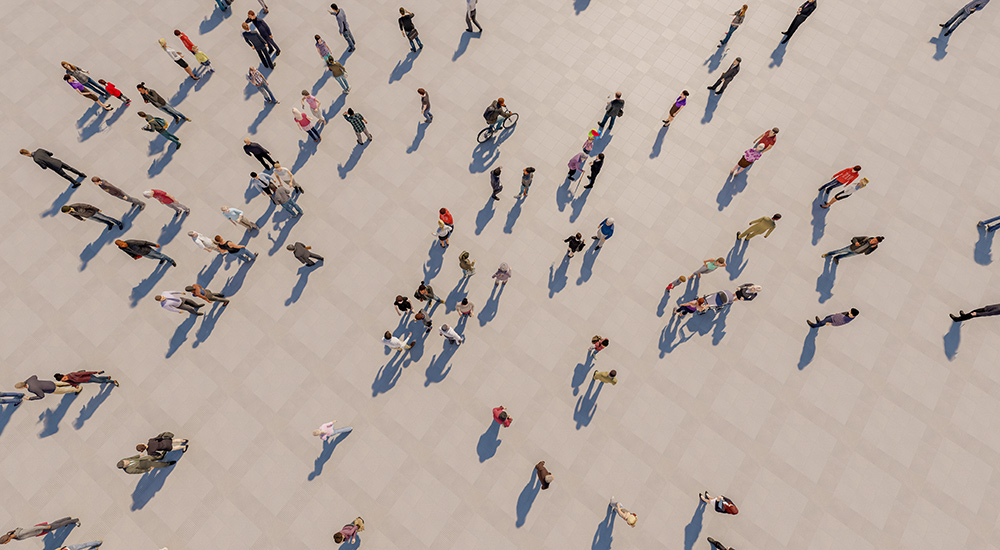 An aerial, top-down photograph of numerous people walking across a light-coloured pavement, casting long distinct shadows. The image captures a diverse flow of pedestrians moving in various directions.