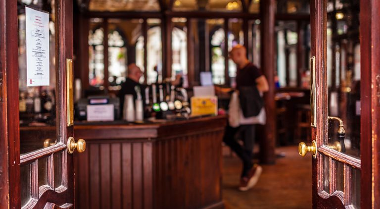 Doors open onto a quiet pub with a wooden interior. A man stands at the bar while another man serves him.