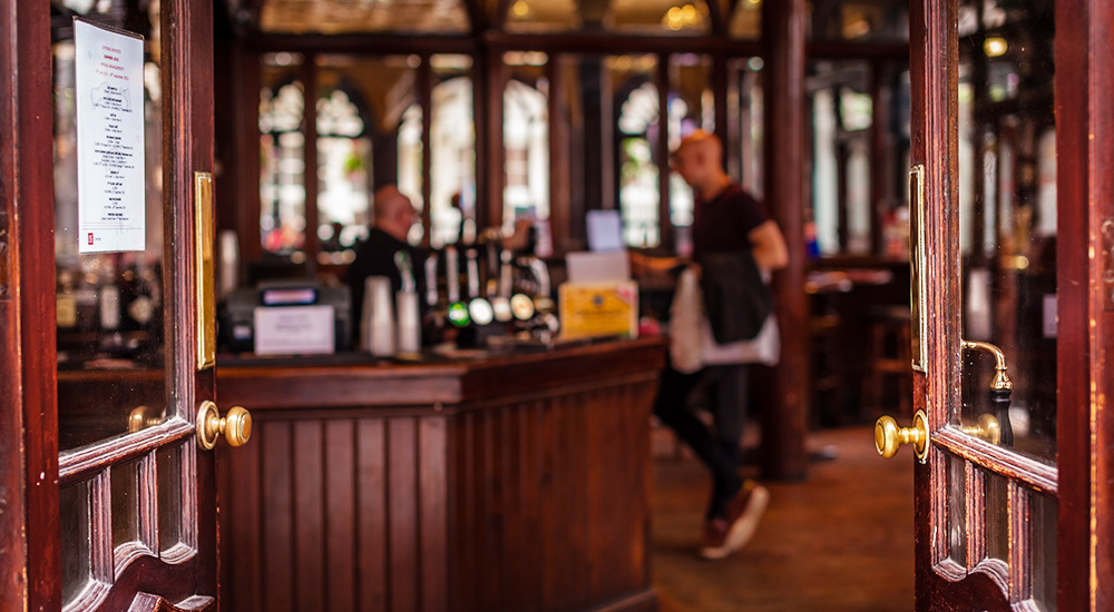 Doors open onto a quiet pub with a wooden interior. A man stands at the bar while another man serves him.
