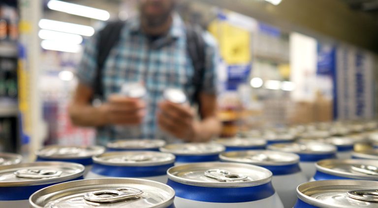 A man in a supermarket holds two cans of beer. In the foreground are many cans of blue and white beer.